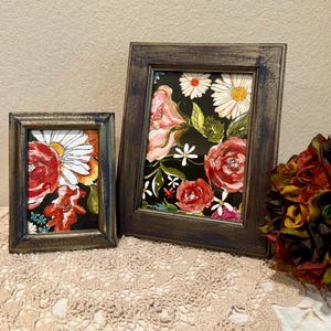 May include: Two framed floral prints with black backgrounds and colorful flowers. The larger frame is dark brown with a distressed finish. The smaller frame is a lighter brown with a gold finish.