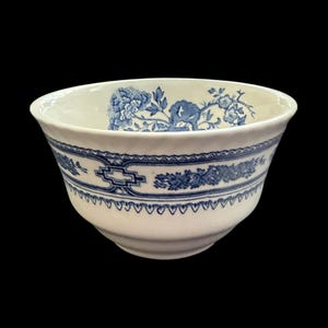 May include: A white ceramic bowl featuring a blue floral pattern around the exterior and interior rim. The design includes a central band with geometric shapes and intricate botanical illustrations of flowers and leaves on a smooth surface.