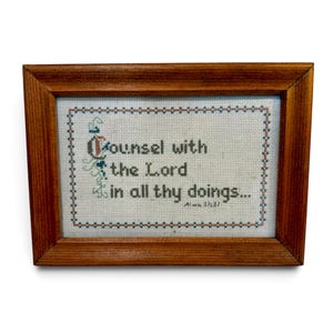 May include: A rectangular wooden frame containing a cross-stitch sampler on white fabric. The text reads "Counsel with the Lord in all thy doings... Alma 37:37" in dark grey thread, surrounded by a decorative border of small blue and red squares.