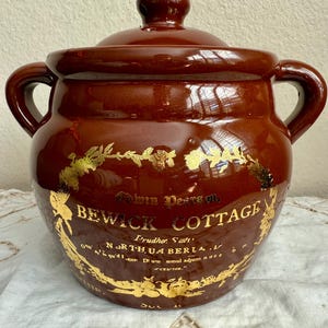 May include: A brown ceramic Bewick Cottage jar with a lid and two handles. Gold lettering and floral designs adorn the jar. The jar is likely used for storage and has a glossy finish.