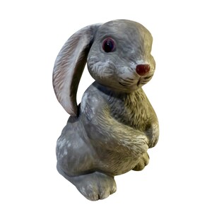 May include: A gray bunny figurine with a seated pose. The rabbit has long ears, red eyes, and a red nose. The figurine is made of a textured material, possibly ceramic or stone, and is suitable for indoor or outdoor decor.