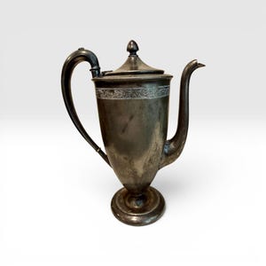 May include: Antique silver-colored coffee pot with a curved handle and spout. The pot has a decorative band around the top and sits on a round base. The lid is topped with a small finial.