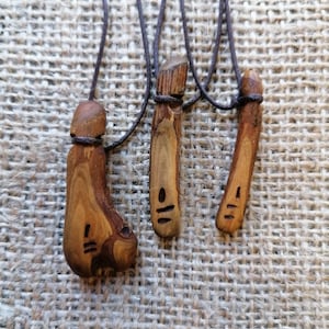 Puede incluir: Tres colgantes de madera con cordones de cuero marrón. Cada colgante tiene un símbolo diferente tallado en él. Los colgantes están sobre un fondo de arpillera marrón.