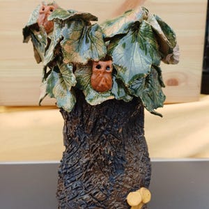Peut inclure: Une sculpture en céramique représentant un tronc d'arbre texturé brun foncé avec un petit champignon jaune à la base. Le sommet est orné de feuilles vertes brillantes et de deux petites figurines de hiboux brunes nichées dans le feuillage.