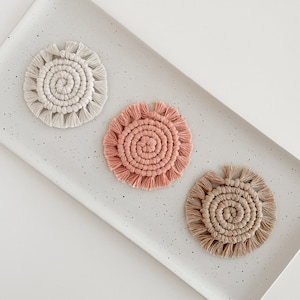 May include: Three handmade macrame coasters in neutral colours: off-white, dusty rose, and tan. Each coaster features a spiral centre with a fringed edge. The coasters are displayed on a speckled grey rectangular tray.