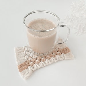 以下が含まれることがあります： 手作りのマクラメコースターの上に置かれた、薄茶色の飲み物が入った透明なガラスマグカップ。 コースターはクリーム色とタン色の幾何学模様で、縁はフリンジ付きです。 マグカップは二重壁のデザインです。