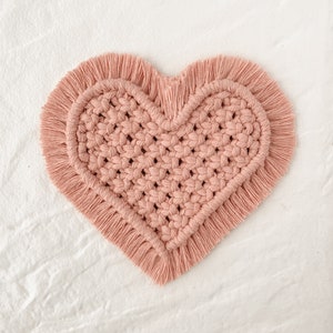 Peut inclure: Un dessous de verre en macramé en forme de cœur, de couleur rose clair. Le dessous de verre présente un centre tissé détaillé et un bord à franges, ajoutant une touche de style bohème. Cet article décoratif est parfait pour ajouter une touche unique à tout décor.