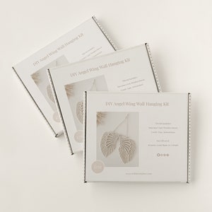 Op de afbeelding: Drie witte dozen met het opschrift "DIY Angel Wing Wall Hanging Kit" zijn diagonaal gerangschikt. Elke doos heeft een macram&eacute; engelenvleugelontwerp. De kit bevat macram&eacute; koord, houten deuvel, koord, tape en instructies.