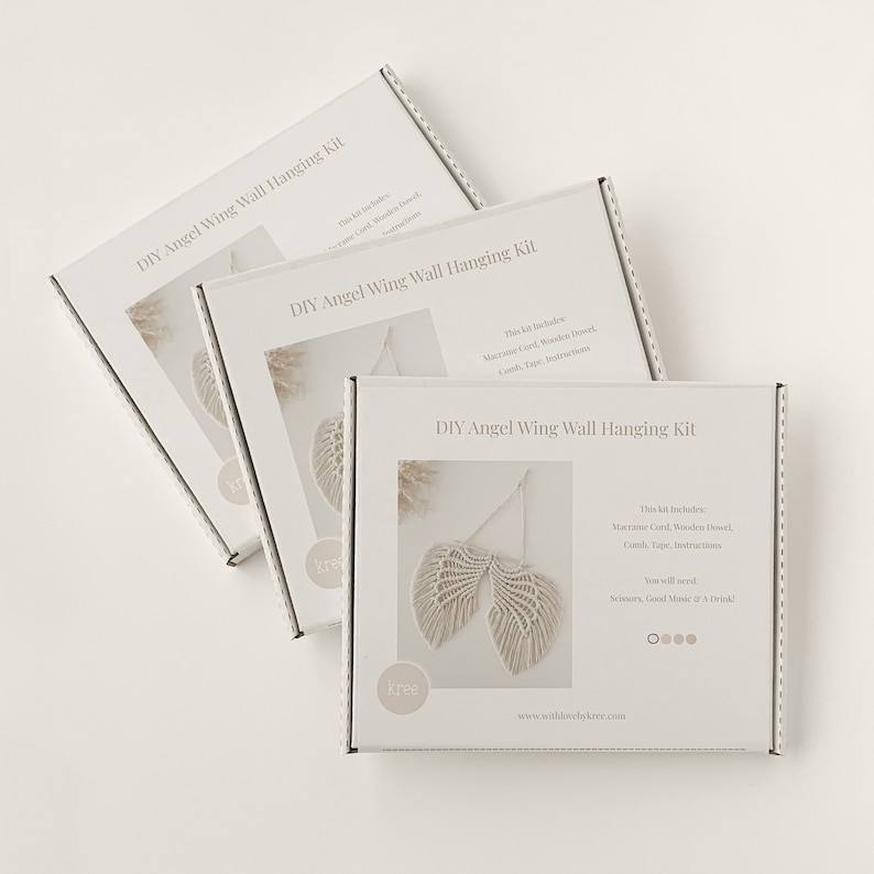 Op de afbeelding: Drie witte dozen met het opschrift "DIY Angel Wing Wall Hanging Kit" zijn diagonaal gerangschikt. Elke doos heeft een macram&eacute; engelenvleugelontwerp. De kit bevat macram&eacute; koord, houten deuvel, koord, tape en instructies.