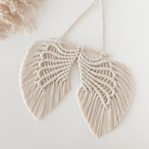 May include: Cream-coloured macrame wall hanging with a feather design. It is suspended from a natural wooden dowel and hanging cord. The macrame has a woven pattern with fringed edges, creating a textured, bohemian aesthetic.