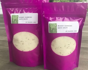 May include: Two purple pouches with white labels that read "Money Drawing Bath Salts by: Shoppe Jinx". Each pouch contains a white bath salt with green herbs.