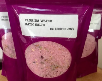 May include: A purple pouch with a white label that reads "FLORIDA WATER BATH SALTS BY: SHOPPE JINX". The pouch contains pink bath salts.