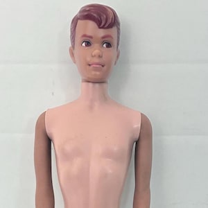 Puede incluir: Una muñeca de plástico con tono de piel claro, cabello moldeado castaño y ojos marrones. La muñeca tiene una complexión delgada, hombros y caderas articulados, y se muestra de pie con los brazos a los lados. Los rasgos faciales incluyen cejas, ojos y labios pintados.