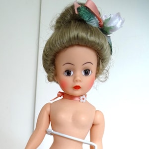 May include: A vintage doll with a painted face, red lips, and rosy cheeks. The doll has blonde hair styled with a decorative bow. It is nude and stands on a white metal stand.