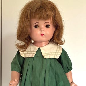Puede incluir: Muñeca vintage con cabello castaño rojizo y un vestido verde con cuello blanco. La muñeca lleva zapatos negros con ribete blanco. La muñeca está sobre un soporte de metal.