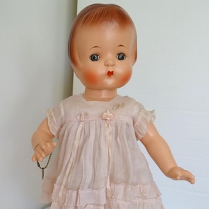 Puede incluir: Una muñeca de porcelana vintage con cabello castaño, ojos azules y un vestido rosa con volantes. La muñeca está de pie sobre una superficie blanca.