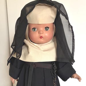May include: A vintage doll dressed as a nun, with a black habit and veil, a cream-coloured wimple, and a rosary. The doll has a painted face with blue eyes and red lips. The doll is standing on a white surface.