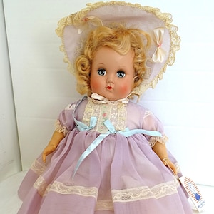 Può includere: Una bambola vintage con capelli biondi ricci che indossa un vestito lavanda con bordino in pizzo e un cappello abbinato. La bambola indossa scarpe bianche e ha gli occhi azzurri.
