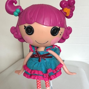 May include: A Lalaloopsy doll with a pink head and hair, large black eyes, and rosy cheeks. The doll wears a turquoise dress with pink trim, red polka dot leggings, and black shoes with pink bows. The doll is standing on a silver stand.