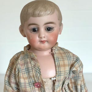 Könnte beinhalten: Eine antike Porzellanpuppe mit bemaltem Gesicht, einschließlich rosiger Wangen und detaillierten Augen. Die Puppe trägt ein kariertes Hemd mit einem rosa Knopf. Das Haar der Puppe ist kurz und ordentlich geschnitten. Eine Sammlerpuppe.