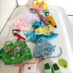 Può includere: Una collezione di accessori per bambole, tra cui un veicolo giallo, ombrelli, tutù, un costume da farfalla, un vestito verde con stampa giungla, una statuina di Babbo Natale, una borsetta, zoccoli verdi e una maschera. Gli articoli sono di vari colori.