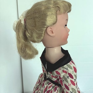 Può includere: Una bambola vintage con capelli biondi raccolti in una coda di cavallo. La bambola ha un incarnato chiaro e indossa un vestito con sfondo bianco e un motivo di foglie rosse e grigie. L'abito ha un colletto nero.