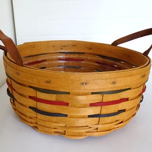 Puede incluir: Una cesta tejida con asas de cuero. La cesta está hecha de madera natural y tiene un diseño multicolor con rayas rojas, azules y verdes.