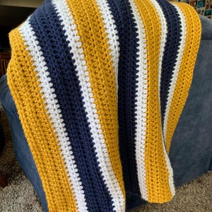 May include: A handmade crochet blanket with vertical stripes in navy blue, mustard yellow, and white. The blanket is draped over a blue sofa, showcasing the textured stitch and color pattern. This cozy throw is perfect for home decor.