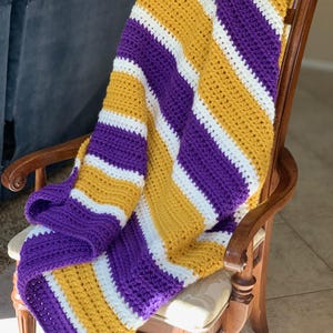 Puede incluir: Una manta de punto de ganchillo con rayas diagonales en morado, blanco y dorado. La manta está colocada sobre una silla de madera.