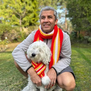 Puede incluir: Un hombre y un perro blanco llevan bufandas de ganchillo a rayas rojas, amarillas y blancas a juego. El hombre lleva una chaqueta gris con cremallera y pantalones cortos. El perro est&aacute; sentado en la hierba. El fondo es un c&eacute;sped verde y &aacute;rboles.
