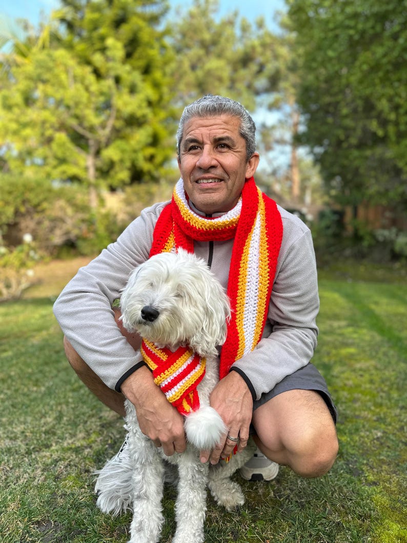 Puede incluir: Un hombre y un perro blanco llevan bufandas de ganchillo a rayas rojas, amarillas y blancas a juego. El hombre lleva una chaqueta gris con cremallera y pantalones cortos. El perro est&aacute; sentado en la hierba. El fondo es un c&eacute;sped verde y &aacute;rboles.