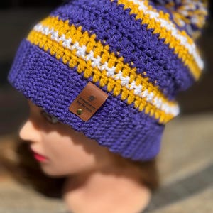 May include: A handmade crochet beanie in purple, gold, and white stripes, topped with a matching pom-pom. The hat features a rolled brim and a small leather tag that reads "Treasures by Teresa".