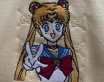 May include: Embroidered patch of Sailor Moon on a light yellow background. The patch features Sailor Moon in her iconic pose with her signature red bow and blue sailor collar. The background is covered in a repeating pattern of the Gucci logo.