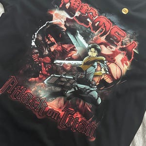 Op de afbeelding: Zwart T-shirt met een grafische print van personages uit de anime Attack on Titan. De print is in een rood-zwart kleurenschema met de tekst "Attack on Titan" in rood.