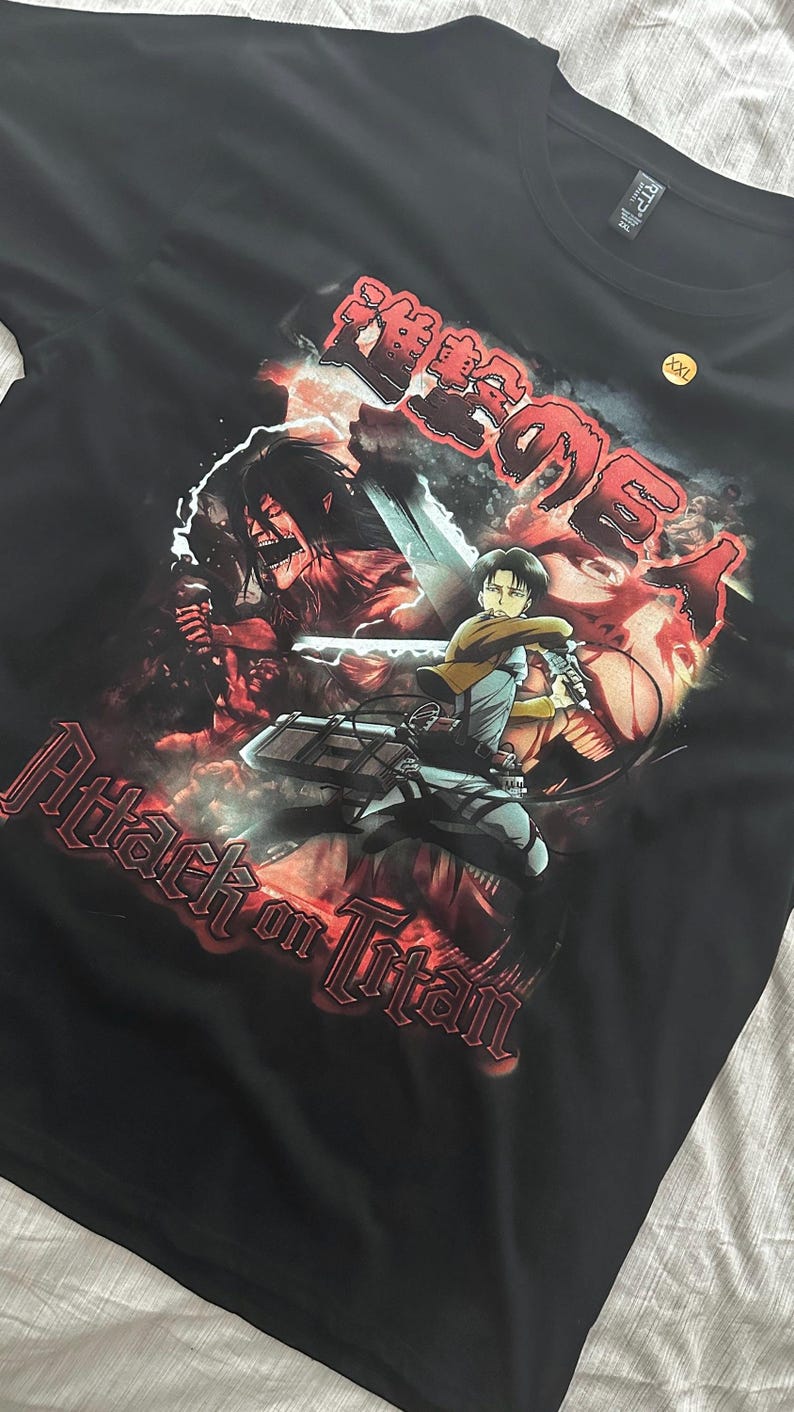 Op de afbeelding: Zwart T-shirt met een grafische print van personages uit de anime Attack on Titan. De print is in een rood-zwart kleurenschema met de tekst "Attack on Titan" in rood.