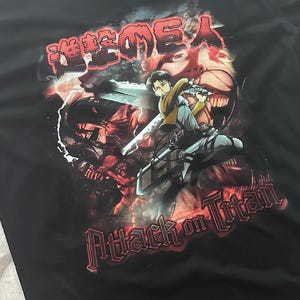Op de afbeelding: Zwart T-shirt met een grafische print van personages uit de anime "Attack on Titan". De grafiek is in een rood-zwart kleurenschema en bevat de Japanse tekst "Shingeki no Kyojin".