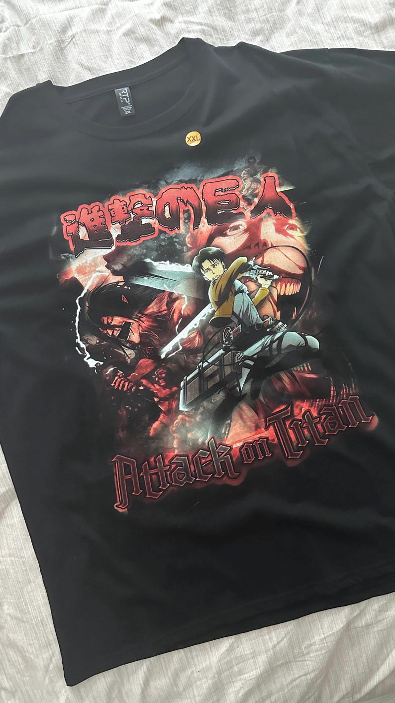 Op de afbeelding: Zwart T-shirt met een grafische print van personages uit de anime "Attack on Titan". De grafiek is in een rood-zwart kleurenschema en bevat de Japanse tekst "Shingeki no Kyojin".