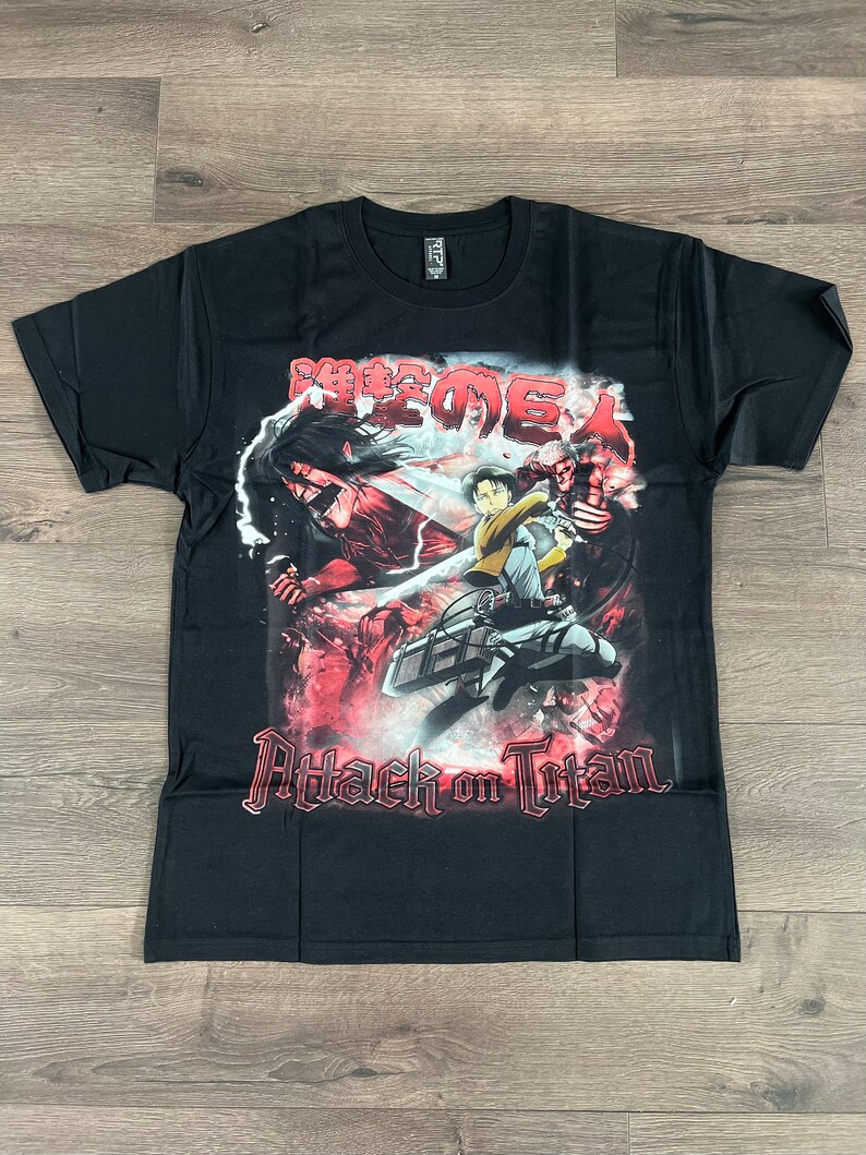 Op de afbeelding: Zwart T-shirt met een grafische print van personages uit de anime Attack on Titan. De grafiek toont een rode en witte achtergrond met de Japanse tekst "Shingeki no Kyojin" en de Engelse tekst "Attack on Titan".