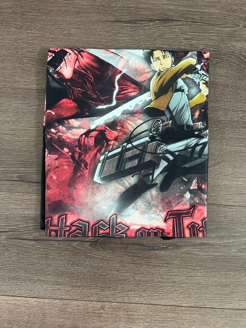 Op de afbeelding: Een zwart T-shirt met een kleurrijke grafische print van een personage uit de anime Attack on Titan. De grafiek toont het personage in actie, met een zwaard en een rood-zwarte achtergrond. De tekst "Attack on Titan" is in zwart gedrukt aan de onderkant van het shirt.
