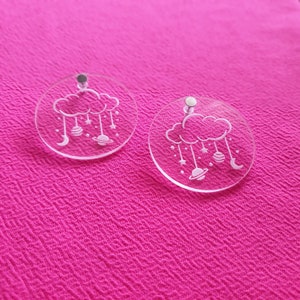 May include: Clear acrylic earrings with a celestial design. Each earring features a round shape with a cloud, stars, planets, and crescent moons. The earrings have silver-colored posts and are displayed on a pink textured surface.