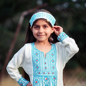 May include: A young girl wearing a white dress with blue and red embroidery. She is wearing a blue and white headband and has long dark hair.