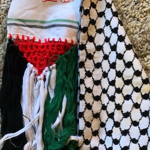 Könnte beinhalten: Ein weißer Stoffscarf mit einem schwarz-weißen Karomuster auf der einen Seite und einem gedruckten Bild der Felsendom-Moschee mit dem Text "عاصمة فلسطين الأبدية" auf der anderen Seite. Der Schal hat Fransen am unteren Rand.