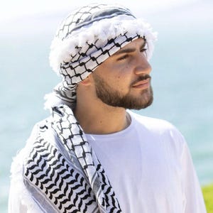 Könnte beinhalten: Ein Mann trägt ein weißes T-Shirt und ein schwarz-weiß gemustertes Kopftuch mit weißem Fransenbesatz.