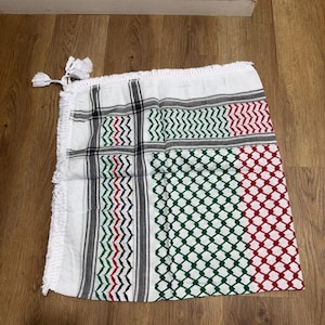 Könnte beinhalten: Weißes Keffiyeh-Tuch mit einem geometrischen Muster in Rot, Grün, Schwarz und Grau. Das Tuch hat einen Fransenrand und eine Quaste an einer Ecke. Das Tuch ist aus leichtem Stoff gefertigt.