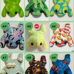 May include: Nine handmade frog plush toys with various fabric patterns. The frogs have black button eyes and are in different colours, including green, blue, and yellow. Some have patterned fabrics with designs like rainbows, moons, and fruit.