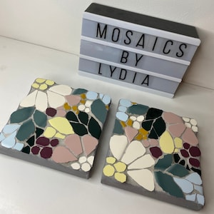 Puede incluir: Dos posavasos cuadrados de hormigón con un diseño floral en mosaico. Los posavasos presentan una mezcla de azulejos rosas, blancos, amarillos, azules, verdes y burdeos.