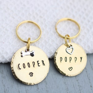 K&ouml;nnte beinhalten: Zwei goldfarbene Hundemarke-Schl&uuml;sselanh&auml;nger mit den eingravierten Namen "Cooper" und "Poppy". Der Schl&uuml;sselanh&auml;nger f&uuml;r Cooper hat einen Knochenanh&auml;nger und einen Pfotenabdruck. Der Schl&uuml;sselanh&auml;nger f&uuml;r Poppy hat einen Herzanh&auml;nger.