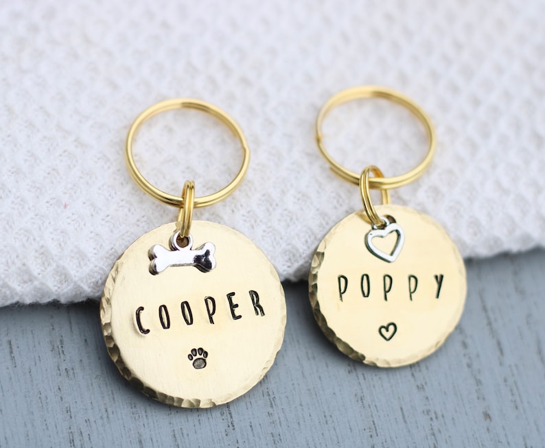 K&ouml;nnte beinhalten: Zwei goldfarbene Hundemarke-Schl&uuml;sselanh&auml;nger mit den eingravierten Namen "Cooper" und "Poppy". Der Schl&uuml;sselanh&auml;nger f&uuml;r Cooper hat einen Knochenanh&auml;nger und einen Pfotenabdruck. Der Schl&uuml;sselanh&auml;nger f&uuml;r Poppy hat einen Herzanh&auml;nger.
