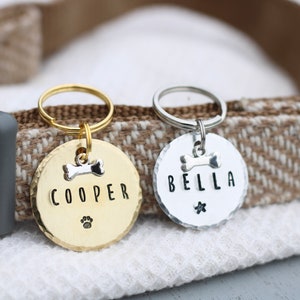 K&ouml;nnte beinhalten: Zwei personalisierte Hundeanh&auml;nger, einer golden und einer silbern, mit den Namen "COOPER" und "BELLA" graviert. Beide Anh&auml;nger haben einen kleinen Knochenanh&auml;nger und einen Pfotenabdruck.