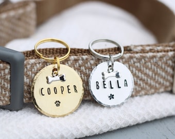 May include: Two personalized dog tags, one gold and one silver, with the names "COOPER" and "BELLA" engraved on them. Both tags feature a small bone charm and a paw print.